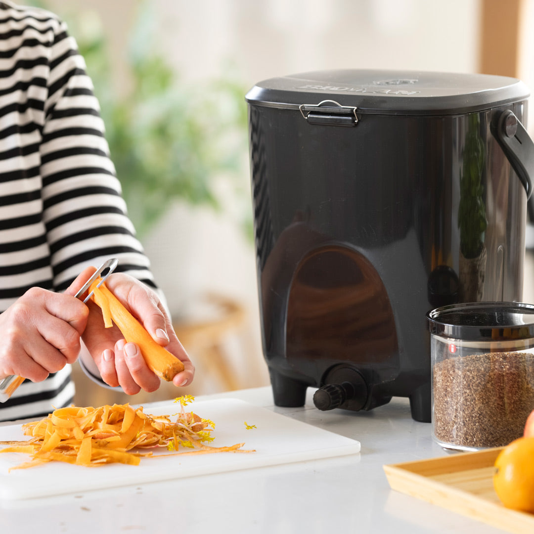 Person peeling carrots next to a black kitchen Bokashi bin on a counter.