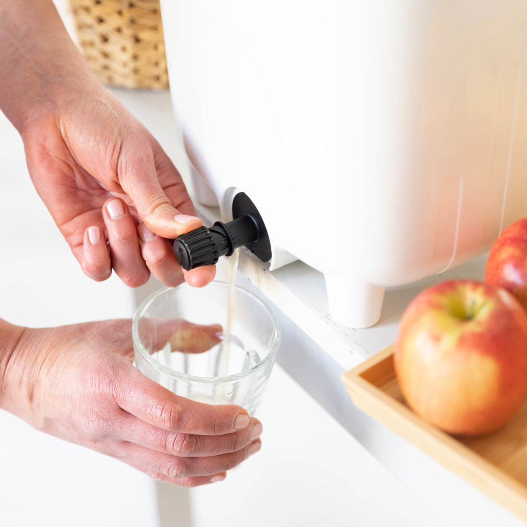 Person pouring liquid from a Bokashi bin into a glass with apples on a tray in the background.
