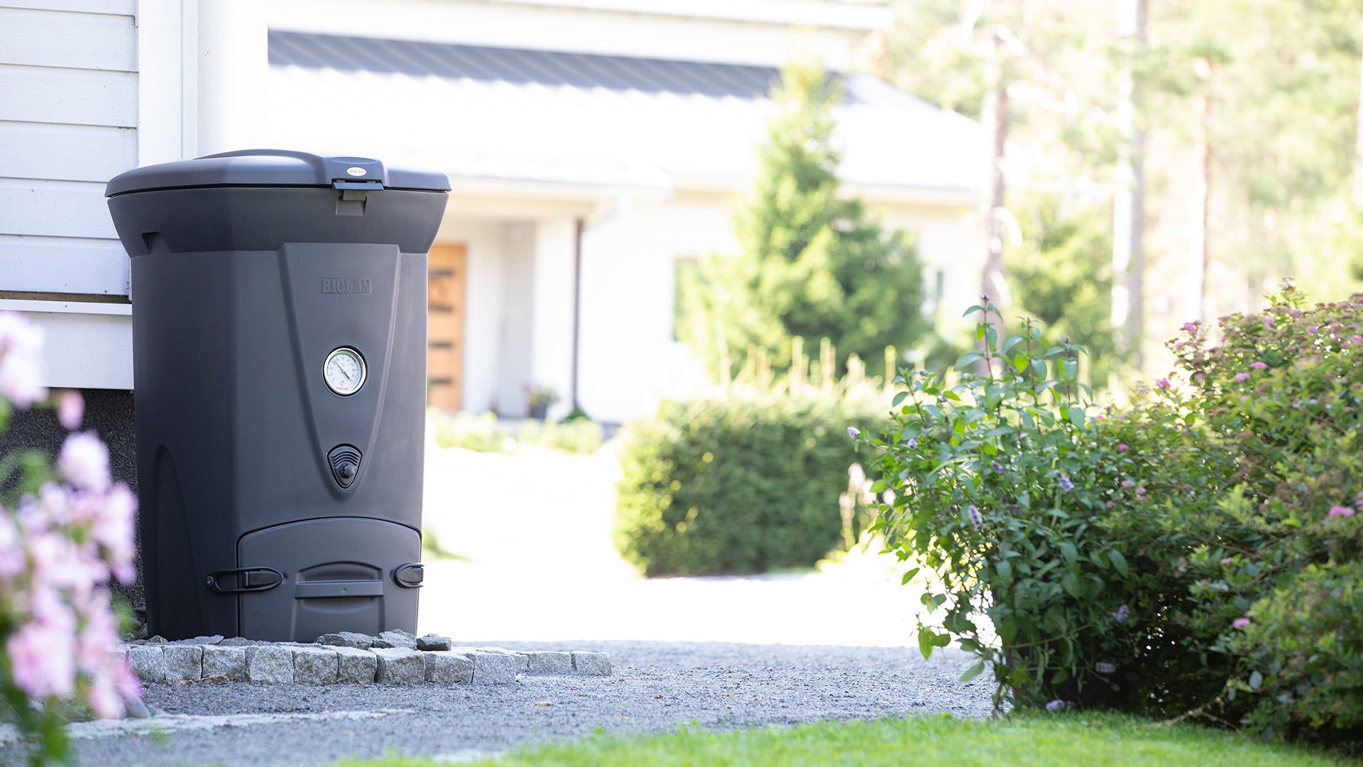 Black compost bin outside a house with a garden in the background